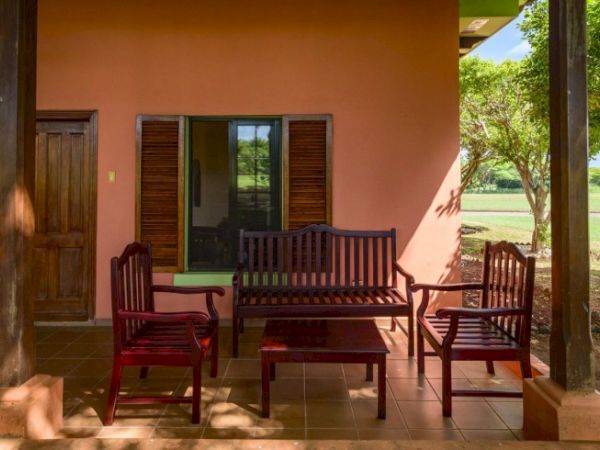 A sunny porch with wooden chairs and a coffee table in front of a peach-colored house.