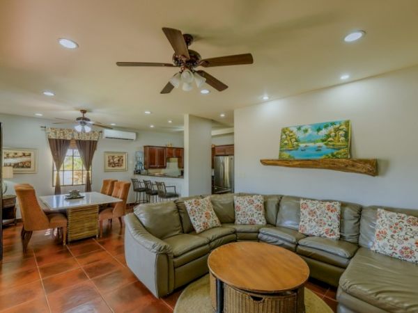 Bright open-plan living room with green sectional sofa, round coffee table, dining area, and kitchen view, plus ceiling fans.