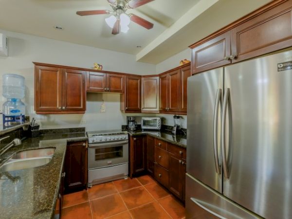 A bright kitchen with dark wood cabinets, stainless refrigerator, stove/oven, granite counters, sink, and a ceiling fan.
