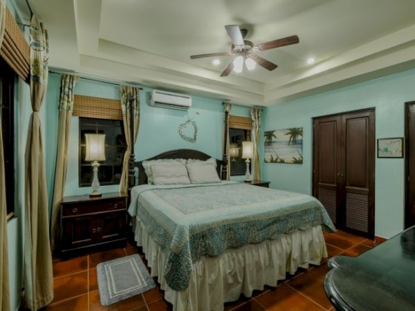 A bright, cozy bedroom with a bed, nightstands, curtains, ceiling fan, and framed decor against blue walls.