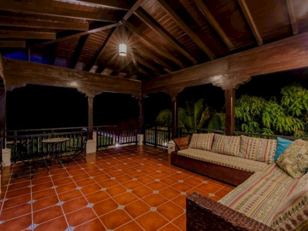 A covered outdoor patio with terracotta tile flooring, wood beams, wicker seating, and string lights surrounding lush greenery.