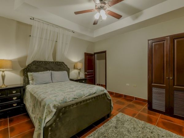 A tidy bedroom with a tufted bed, bedside tables, a ceiling fan, and tall wooden doors and closets, ending in a quiet, warm space.