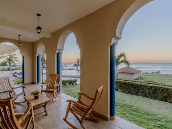 An arched seaside patio with wooden chairs and potted plants, overlooking the ocean at sunset.