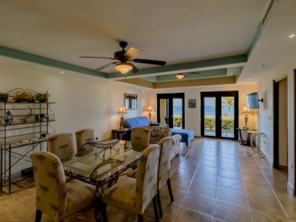 A bright tropical living/dining room with ceiling fan, sofas, upholstered chairs, and large doors showing an ocean view.