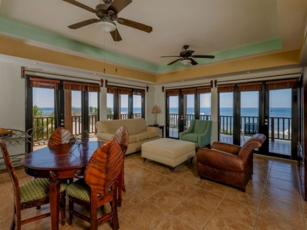 A sunny living room with sofas and chairs, ceiling fans, and large doors opening to an ocean view.