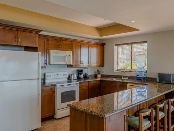 A bright kitchen with wooden cabinets, a refrigerator, stove, microwave, coffee maker, and a granite island by a window.
