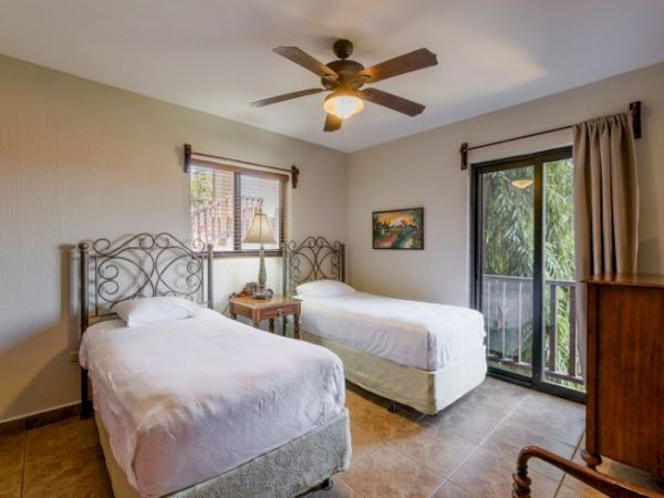 A bright bedroom with two neatly made beds, a ceiling fan, and large windows with curtains overlooking greenery.