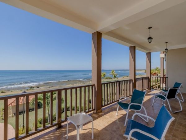 Balcony patio with four blue lounge chairs overlooking a calm ocean and sandy beach under a clear sky.