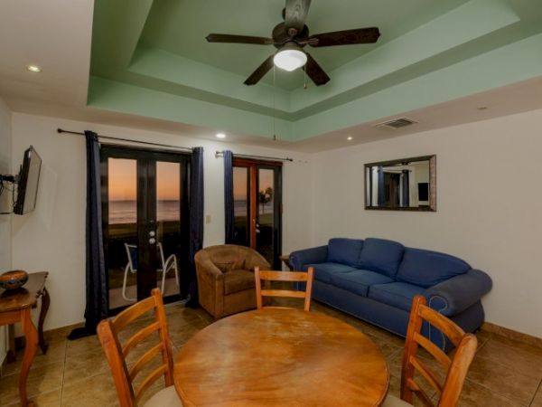A bright living room with a blue sofa, round wooden table, chairs, ceiling fan, and sliding doors showing a sunset ocean view.