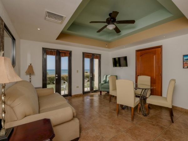 A bright living room with beige sofas, dining chairs and table, ocean-view doors, and a ceiling fan.