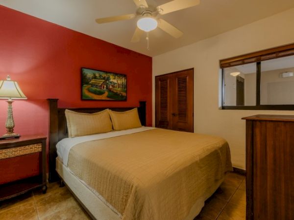 A cozy bedroom with a large bed, red accent wall, ceiling fan, bedside lamp, and wooden dresser/counter.