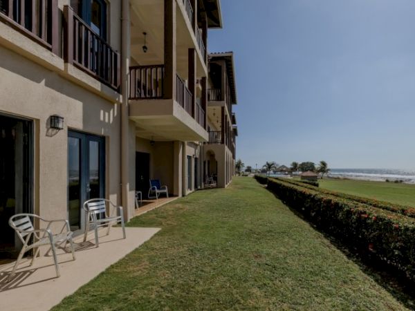 A seaside hotel corridor with balconies, outdoor patio chairs, trimmed grass, and a hedge beside the ocean, under a blue sky.