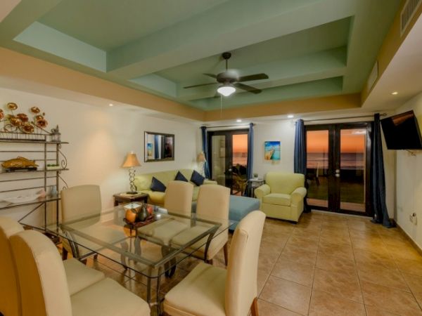 A bright living room with a glass dining table, white chairs, sofas, a ceiling fan, and sliding doors leading outside.