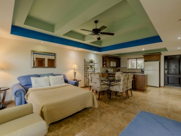 A bright living and dining area with tiled floors, blue accents, a ceiling fan, sofas, and a table set with chairs.
