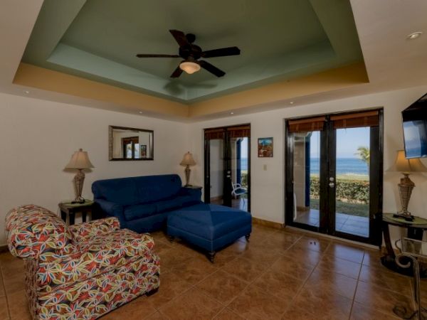 A bright living room with patterned armchairs, a blue sofa, ceiling fan, lamps, TV, and large glass doors opening to a view.