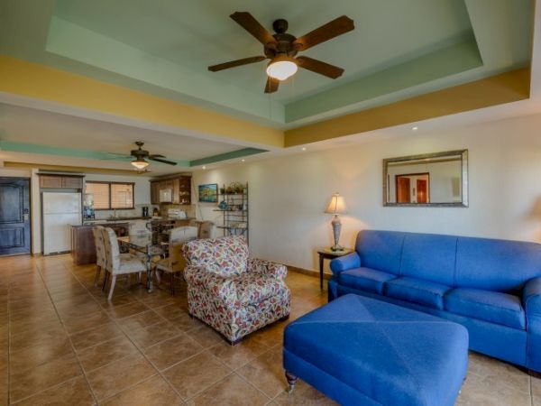 A bright living room with a blue sofa and ottoman, tiled floors, ceiling fans, and an open kitchen with chairs and appliances.