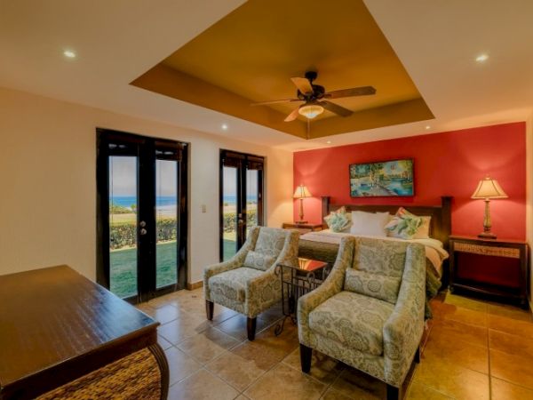A bright bedroom with a ceiling fan, patterned armchairs, a neatly made bed, and large doors opening to an outdoor view.