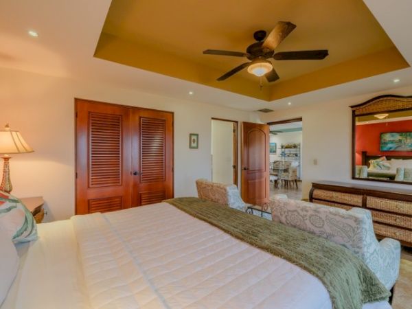 A bright bedroom with a large bed, ceiling fan, wooden doors, and framed wall art.