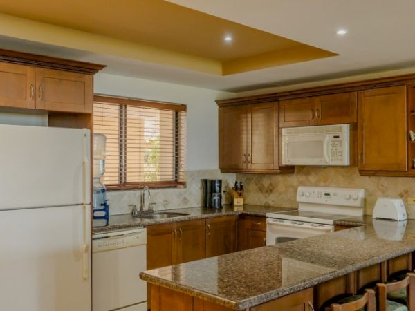 A bright kitchen with wooden cabinets, granite counters, a refrigerator, microwave, stove, and sink under a window.