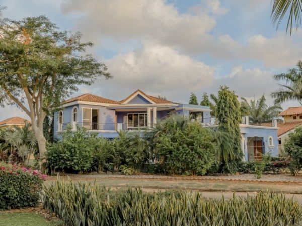 A large house with a red-tile roof, surrounded by lush green landscaping, gardens, and trees under a partly cloudy sky.