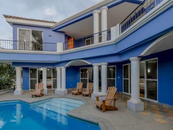 Blue-and-white courtyard of a house with a swimming pool, arched veranda, and wooden lounge chairs.