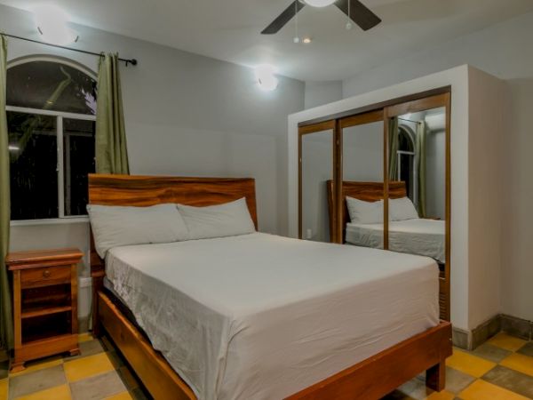 A neatly made bedroom with a wooden bed, bedside table, curtains, mirror wardrobe doors, and a ceiling fan with lights.