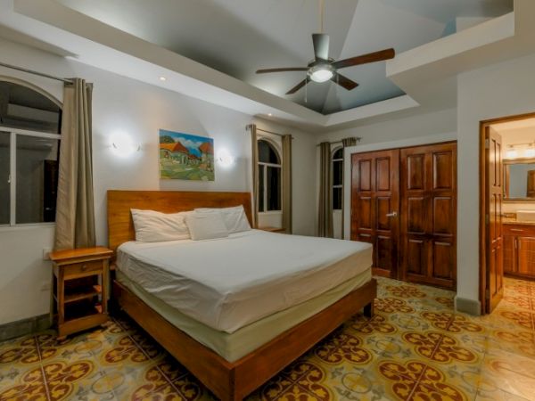 A bright bedroom with a wooden bed, patterned tile floor, ceiling fan, and warm lighting.