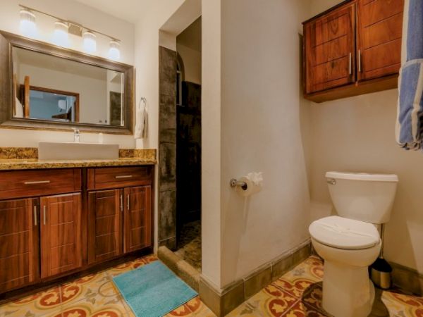 A spacious bathroom with a wooden vanity, large mirror, tiled floor, towel, and a toilet with a small rug in front.