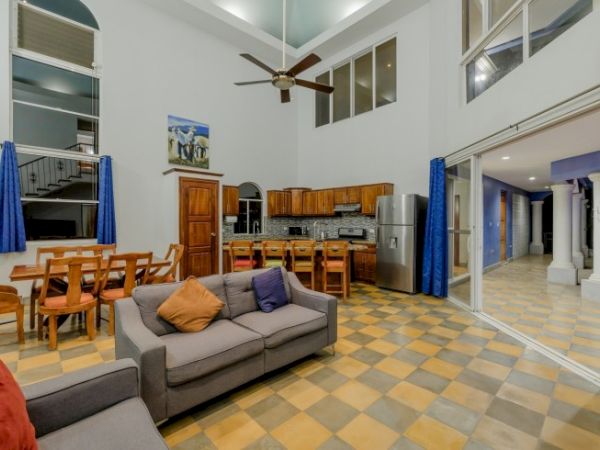Bright open-plan living room and kitchen with a gray sofa, dining table, blue curtains, and stainless appliances.