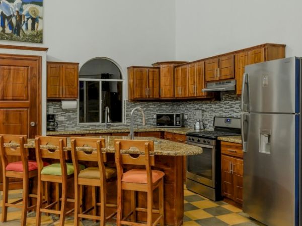 A spacious wood-cabinet kitchen with island and stone countertop, chairs, stove, sink, and stainless steel refrigerator.