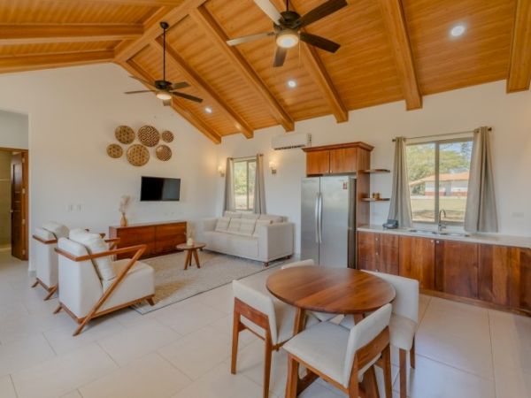 A bright open-plan living and kitchen area with wooden ceiling, sofas, and a dining table, plus stainless appliances and large windows, .