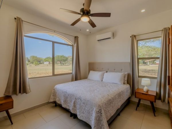 A bright bedroom with a ceiling fan, double bed, large windows with curtains, and an outdoor view.