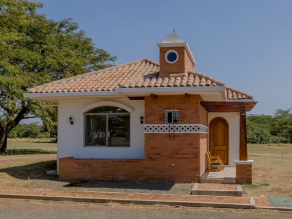 A small brick-and-stucco house with a tiled roof, arched window, wooden door, and a front porch by green trees.
