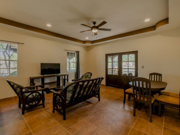 Living room with beige walls, ceiling fan, dark wood sofa and chairs, TV on a stand, and a dining set by glass doors.