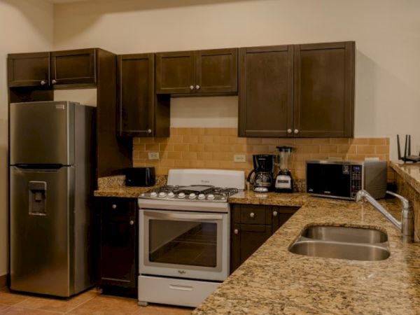 A compact modern kitchen with dark cabinets, refrigerator, oven/stove, granite counters, coffee setup, and a stainless sink.