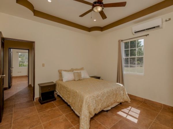 A bright bedroom with a bed, ceiling fan, window with curtains, tiled floor, and a wall air conditioner.