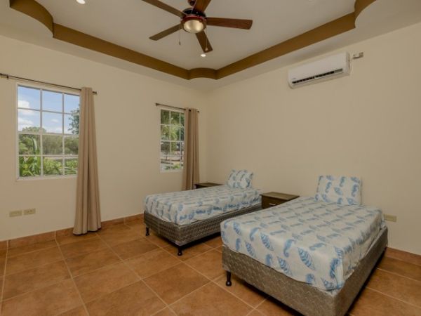 A bright bedroom with two twin beds, blue-pattern bedding, ceiling fan, air conditioner, two windows, and beige curtains.