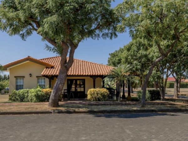 A small house with a terracotta-tiled roof and shaded patio, surrounded by tall trees and a paved driveway.