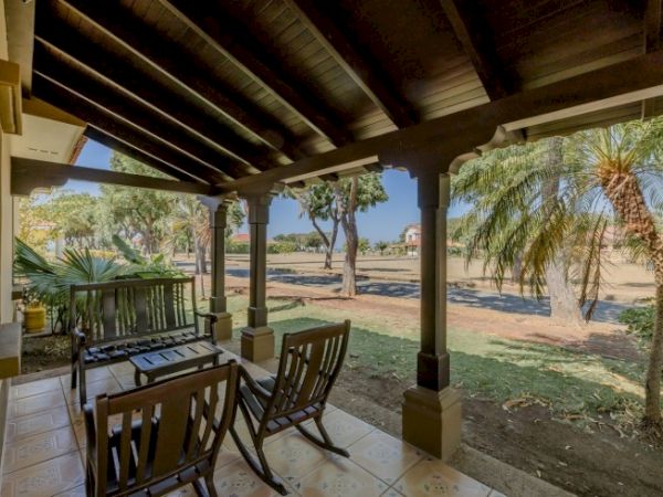 A covered porch with wooden chairs and railings overlooks a sunny yard with palm trees and a road beyond, in warm weather.