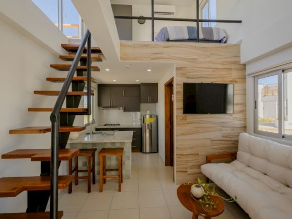 Bright modern interior with a wooden spiral staircase, compact kitchen, flat-screen TV, and cream sofa in an open-plan space.