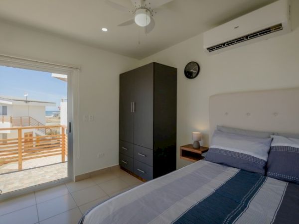 A bright bedroom with a bed, dark wardrobe, bedside table, and ceiling fan, plus a door opening to a balcony.