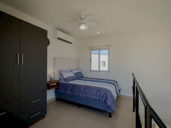 A bright bedroom with a bed, dark wardrobe, ceiling fan, air conditioner, and a window letting in daylight.