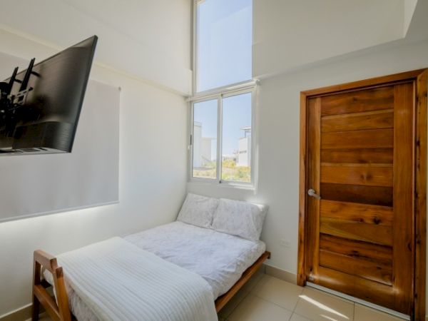 A small bright bedroom with a single bed, white bedding, a large window, and a wooden door.