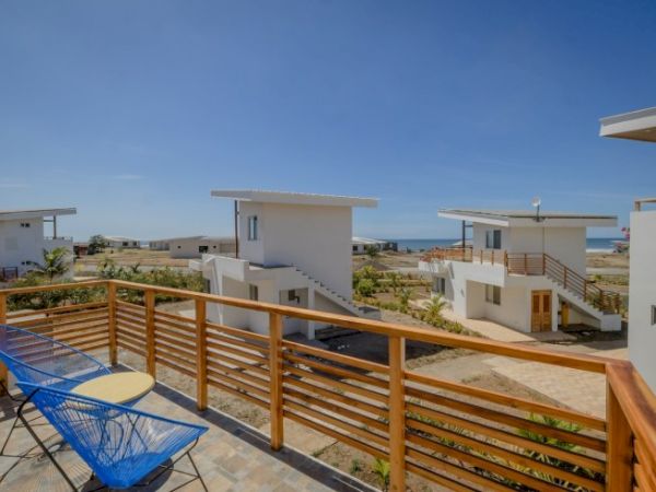 A beachfront resort balcony with wooden railing, a blue lounge chair, and modern white villas overlooking the sea.
