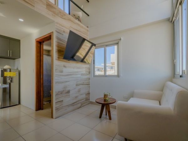 A modern living room with a white sofa, small round table with plant, large window, tiled floor, and a wall-mounted TV, beautifully staged.