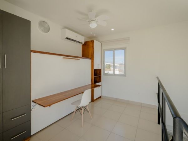 A modern bright room with a built-in wooden desk/shelves, white chair, window view, tiled floor, and ceiling fan and AC.
