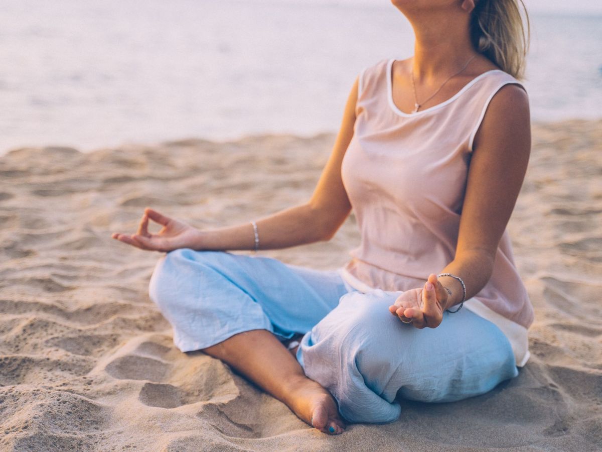 A woman sits cross-legged on a sandy beach, meditating with eyes closed as the waves roll in behind her.