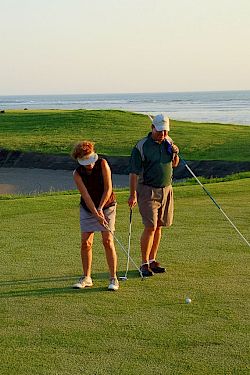 A group of golfers practices on a seaside green, chatting while holding clubs as waves roll in the distance.