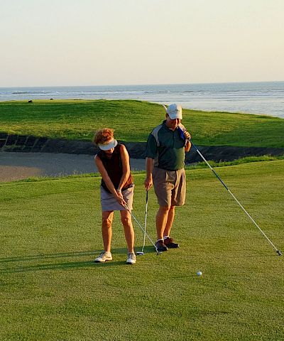 A group of golfers practices on a seaside green, chatting while holding clubs as waves roll in the distance.