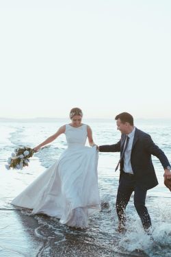 A bride in a white gown wades into the surf with a groom in a suit holding her bouquet, on a windy beach shore.